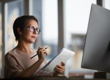 Woman looking at her computer while hand-writing on a notepad
