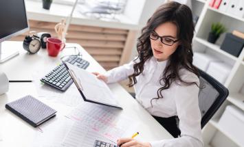 Person with long hair working at a desk with a calculator and computer